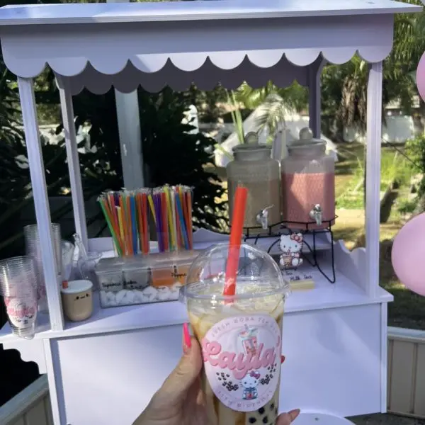 A person holding a decorated milkshake cup in front of a small outdoor Elegant White Snack or Candy Cart Rental with jars of toppings and drinks.