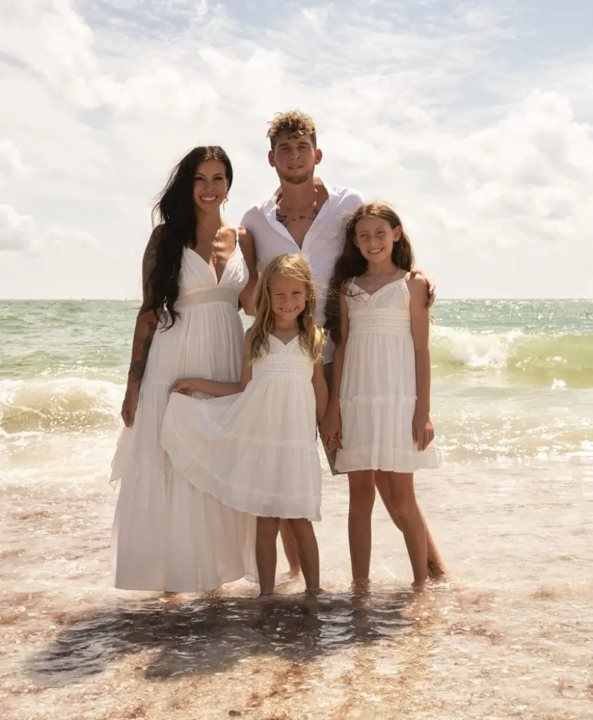 A family of four, dressed in white, standing in shallow ocean water under a sunny sky. Two adults and two children smiling towards the camera.