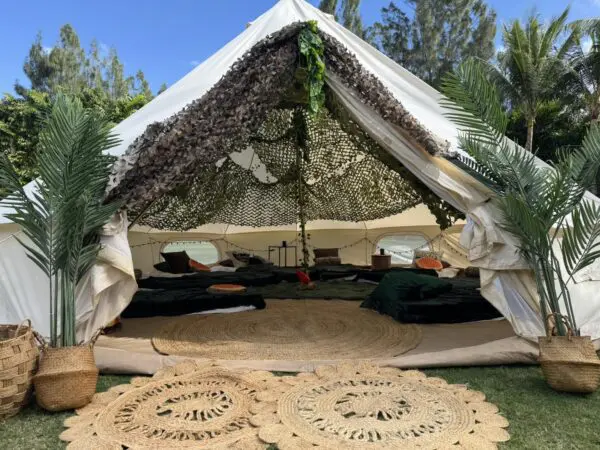 Discover the ultimate Camping Party experience with a stunning open-front white tent featuring a unique camouflage netting roof. This large outdoor setup is beautifully decorated with cozy baskets and vibrant rugs, creating an inviting atmosphere perfect for any celebration. Nestled on lush green grass and surrounded by towering trees, this enchanting setting offers an ideal blend of luxury and nature, making it the perfect backdrop for memorable gatherings in the great outdoors. Ideal for camping parties or rustic-themed events, this picturesque scene will captivate your guests and provide an unforgettable ambiance under the open sky.