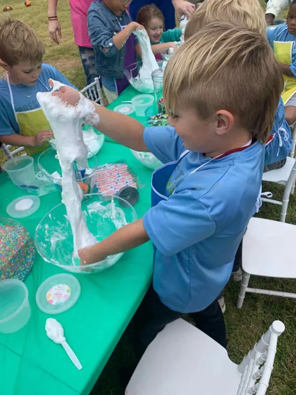 A young boy in a blue t-shirt engages with a slime-making activity at a green table during the 2024 Ultimate Sleepover, surrounded by other children outdoors.