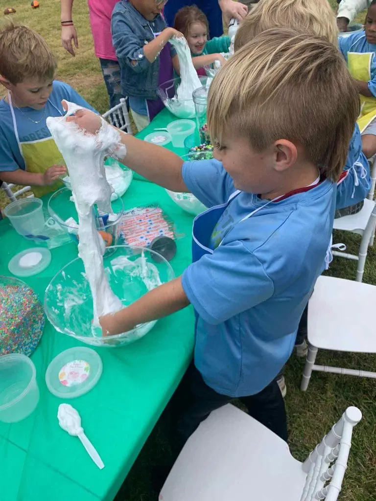 A young boy in a blue t-shirt engages with a slime-making activity at a green table during the 2024 Ultimate Sleepover, surrounded by other children outdoors.