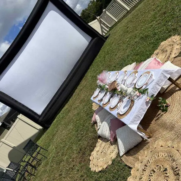 Outdoor movie setup with a large screen and a decorated table with plates and cushions on the grass for the ultimate night.