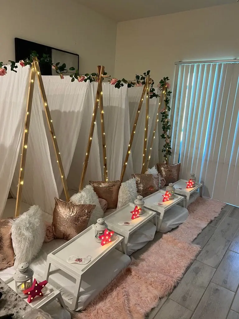 Indoor picnic setup with glam-themed party tents adorned with fairy lights and roses, floor cushions, and trays with candles, against a backdrop of vertical blinds.