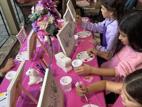 A group of enthusiastic children is seated around a vibrantly decorated table, fully engaged in painting intricate butterfly outlines on their canvases. Their creativity is as dazzling and vivid as the atmosphere of a Movie Night Extravaganza. The table itself bursts with charm, adorned in a delightful pink cloth and sprinkled with whimsical flowers, enhancing the enchanting ambiance of this artistic gathering. Keywords: children painting butterflies, creative art session for kids, whimsical table setting with flowers.