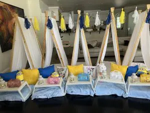 A cozy room setup featuring four small tents with pillows, blankets, lanterns, and Minion toys, as if straight from an AUTO-DRAFT layout. Blue and yellow tassel garlands hang above, with a sofa and kitchen area visible in the background.