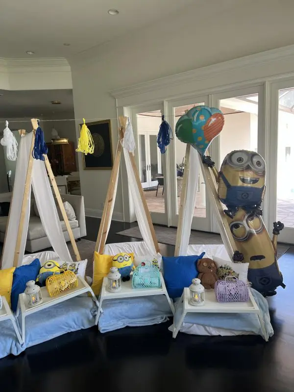 Three indoor tents with pillows and blankets, each with a tray holding a lantern, teddy bear, and gift bag. An AUTO-DRAFT balloon stands near the rightmost tent, next to the Minion-themed balloon.