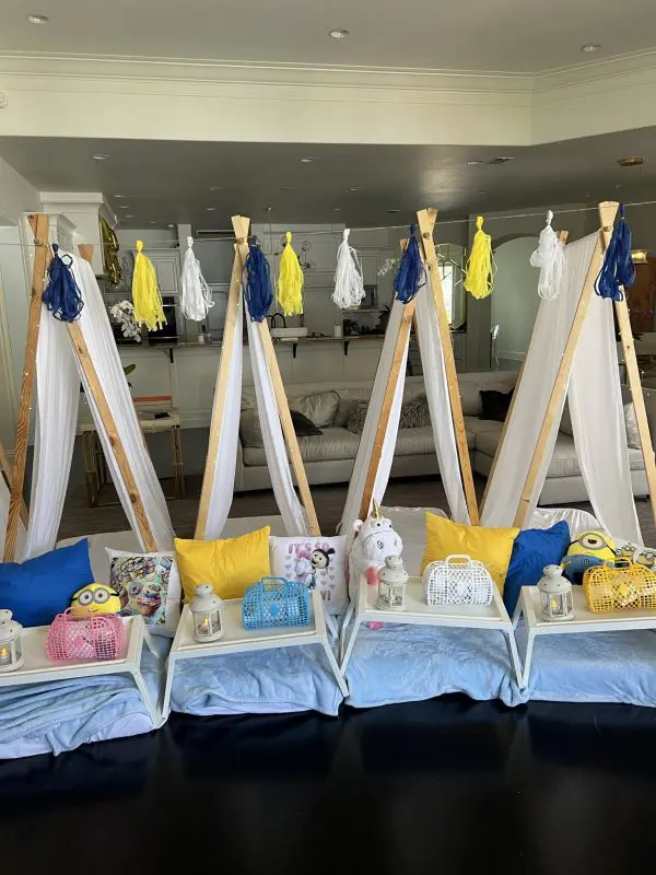 Four indoor teepees arranged with colorful pillows and lanterns are set up in a living room, adorned with yellow, blue, and white decorative tassels hanging above them. The cozy setting feels like it was designed on auto-draft but brings an effortless charm to the space.