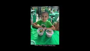 A young girl in a green shirt and headband sits at a table with two bowls of colorful slime at JoJo's Shake Bar. With a smile on her face, she looks at the camera, as if to say thank you for this fun experience.