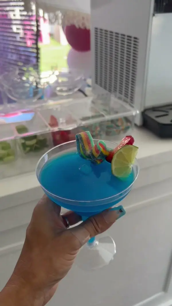 A hand holding a blue cocktail in a martini glass, garnished with colorful candy, a lime wedge, and a strawberry, creates an air of luxury reminiscent of our Luxury Soft Play Packages in front of a countertop with various ingredients in clear bins.