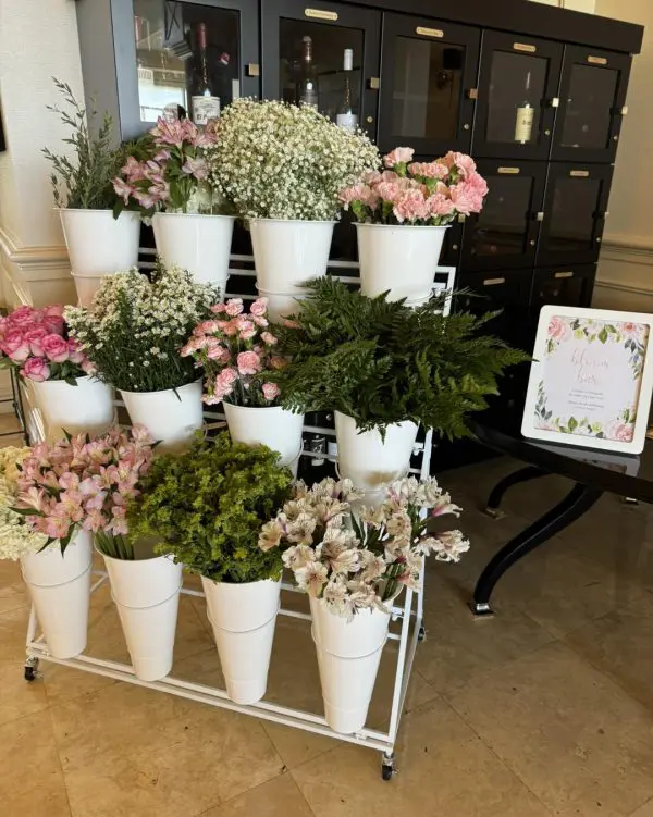 A captivating flower cart brimming with elegance showcases a stunning floral display, featuring white ceramic vases overflowing with delicate pink and white blooms. The arrangement includes enchanting pink roses and airy sprigs of baby's breath, artfully presented on a sophisticated multi-tiered metal stand. A charming decorative sign nearby invites passersby with the words "Flowers for You," adding an inviting touch to the scene. In the background, a sleek black cabinet with multiple compartments complements the vibrant floral ensemble, offering an enticing backdrop to this picturesque flower display. Perfect for anyone seeking inspiration in floral arrangements or lovers of exquisite nature-themed decor!