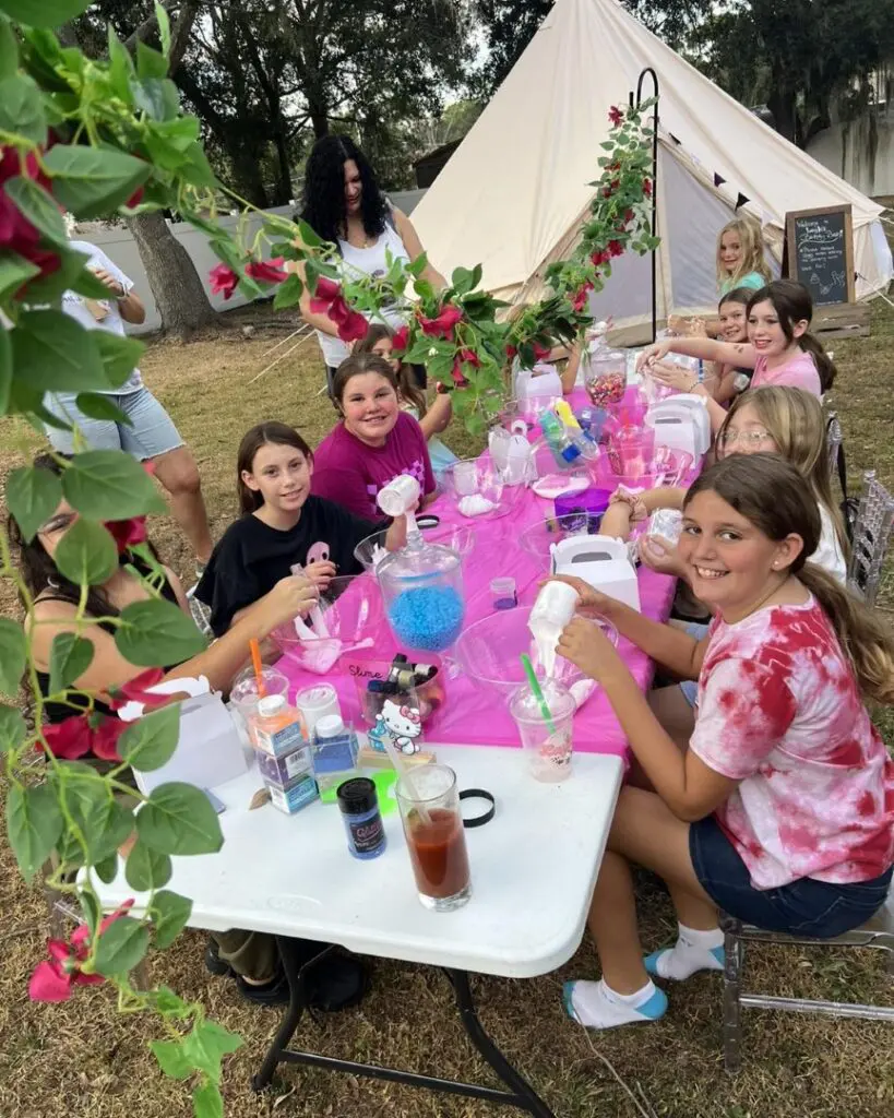 A joyful group of children eagerly engages in arts and crafts at a vibrant outdoor setting, gathered around a long table adorned with a cheerful pink cloth. Underneath the canopy of a cozy tent in the background, they immerse themselves in creative projects using an array of colorful materials. Nearby, an encouraging woman stands by to assist and inspire young imaginations. The picturesque scene is beautifully framed by blooming flowers that add an extra touch of nature's charm, enhancing this delightful outdoor crafting experience. Perfect for family-friendly activities and children's creativity inspiration!.