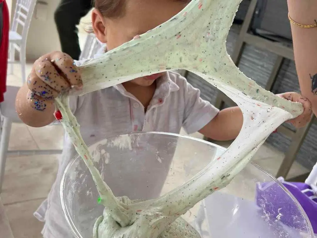 A child playfully stretches a vibrant, gooey slime over a large mixing bowl, enchanting the senses with its bright and squishy texture. Tiny hands sparkled with shimmering blue glitter, adding a touch of magical sparkle that catches the light beautifully. Explore the joy of sensory play as this little one indulges in hands-on creativity and messy fun! Perfect for those seeking engaging kids' activities with colorful crafts and glittery DIY experiments.