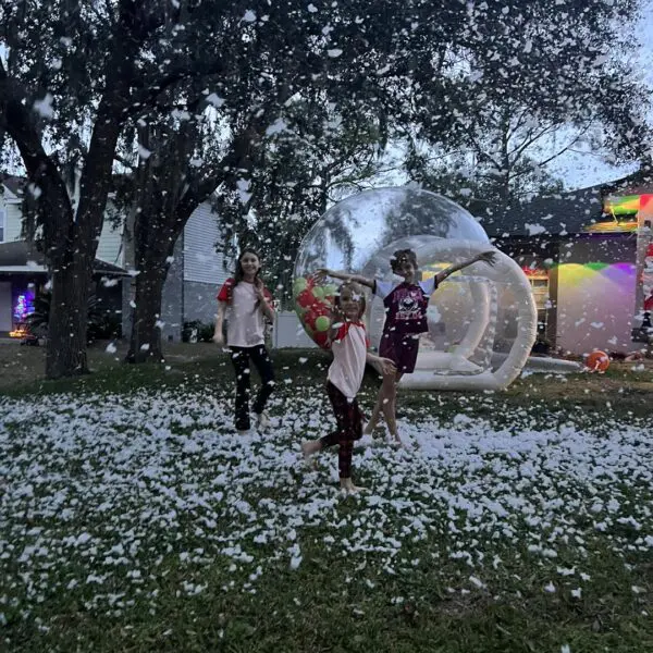 Three joyful children frolic in a winter wonderland of artificial snow, thanks to a rented snow machine transforming their backyard into a festive scene. Behind them stands an impressive inflatable transparent dome, creating a surreal and magical atmosphere. The vibrant colors of the house in the background add to the lively and enchanting setting, making it perfect for capturing memorable moments and bringing holiday cheer right to your home. Ideal for winter-themed parties or celebrations even in warmer climates!