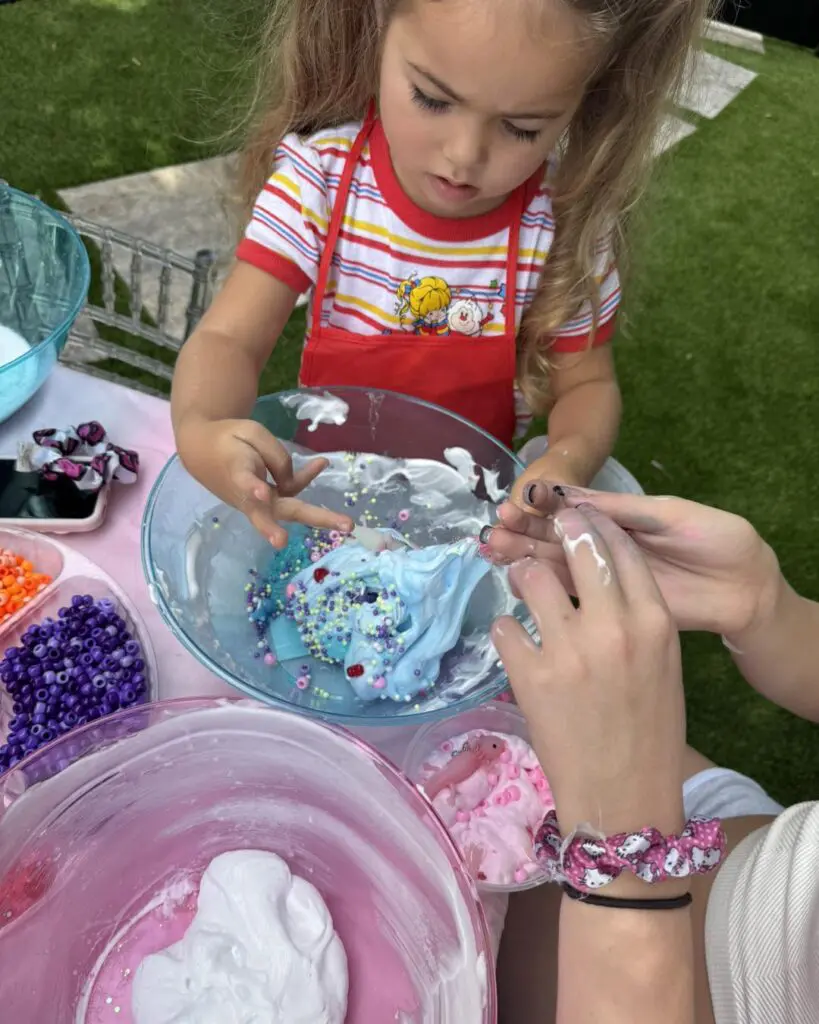 Young child in a vibrant striped shirt and bright red apron joyfully explores the sensory delight of slime play at a creative activity table. The tabletop is an explosion of color, adorned with an assortment of bowls brimming with multicolored beads and fluffy shaving cream, perfect for stimulating imaginative play. This hands-on experience not only captivates curiosity but also enhances motor skills and creativity. Perfect for parents seeking engaging activities for kids involving tactile learning and fun.