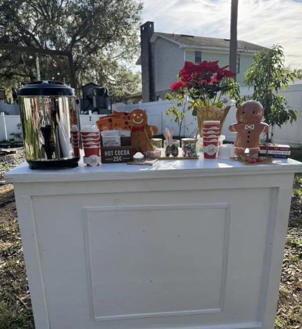 Delight in a picturesque outdoor hot cocoa stand that captures the essence of a winter wonderland. At the heart of this enchanting setup is a gleaming silver urn, surrounded by whimsical gingerbread decorations and vibrant poinsettias. Packs of cocoa mix and classic candy canes are artfully arranged on the pristine white table, inviting all to indulge in some holiday cheer. The magic of Snow Machine Rental's swirling snowflakes dances around this festive display, transforming it into an idyllic scene straight from your winter dreams. Perfect for those seeking heartwarming seasonal charm and Instagram-worthy moments, this setup promises to enthrall both locals and tourists with its cozy, captivating allure.