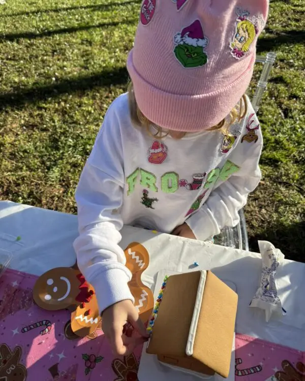 A joyous child in colorful festive attire lovingly decorates a charming gingerbread house at a vibrant outdoor table. Nearby, the Snow Machine Rental creates a magical winter wonderland ambiance with enchanting, gentle snow flurries swirling around. Perfect image capturing holiday cheer and creativity in a festive setting!