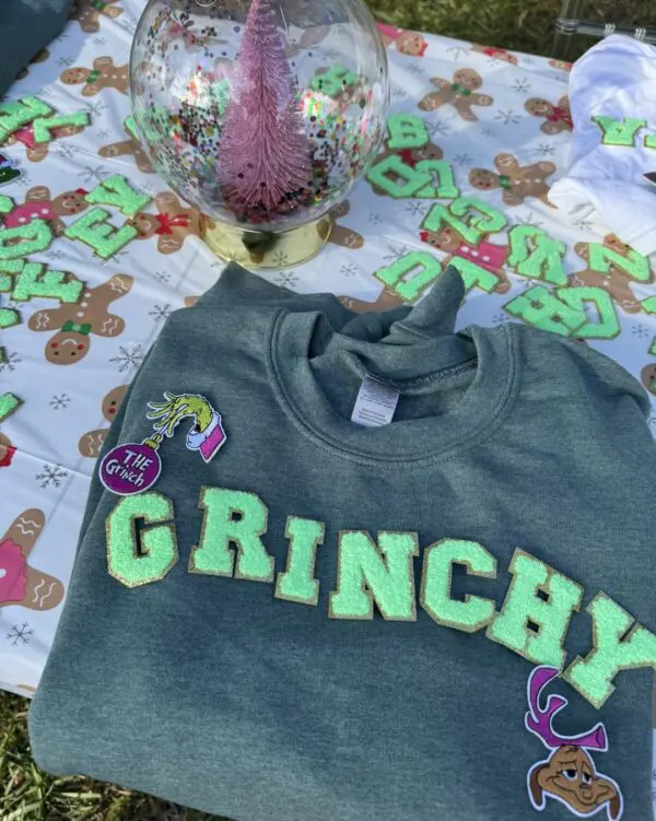 A vibrant green sweatshirt emblazoned with bold "GRINCHY" lettering lies prominently on a table, perfectly capturing the playful essence of holiday cheer. Nestled beside this cozy attire is a dazzling pink glittery tree ornament, exuding festive sparkle and charm. Complementing the scene is whimsical gingerbread-themed decor, adding a touch of warmth and sweetness to your holiday aesthetic. Elevate your seasonal celebration with this enchanting setup that's ideal for any holiday party. Enhance the wintry ambiance even further with the magical touch of a Snow Machine Rental, creating an unforgettable festive experience perfect for sharing on social media!