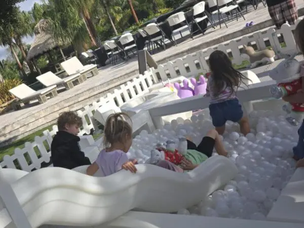 Children delight in a vibrant white ball pit, enclosed within a secure fence, offering endless fun and laughter. Nearby, the Movie Concession Stand Rental transforms the area into a cinema-inspired setting, complete with cozy lounge chairs and stylish tables. This playful scene captures the essence of a theater lobby experience, perfect for event rentals or family gatherings. Immerse yourself in this entertaining atmosphere where movie magic meets playtime excitement!.