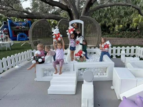 Children immerse themselves in imaginative outdoor play on versatile modular furniture, surrounded by an array of plush stuffed toys. The playful scene, set against a picturesque backdrop of tall trees and a charming white picket fence, is bursting with excitement. An eye-catching inflatable structure adds a layer of fun and adventure, while a trendy Movie Concession Stand Rental provides a delightful touch, offering snacks and treats to fuel their creativity. This whimsical setup creates the perfect environment for kids' imaginative adventures, making it an ideal theme for outdoor events or family gatherings.