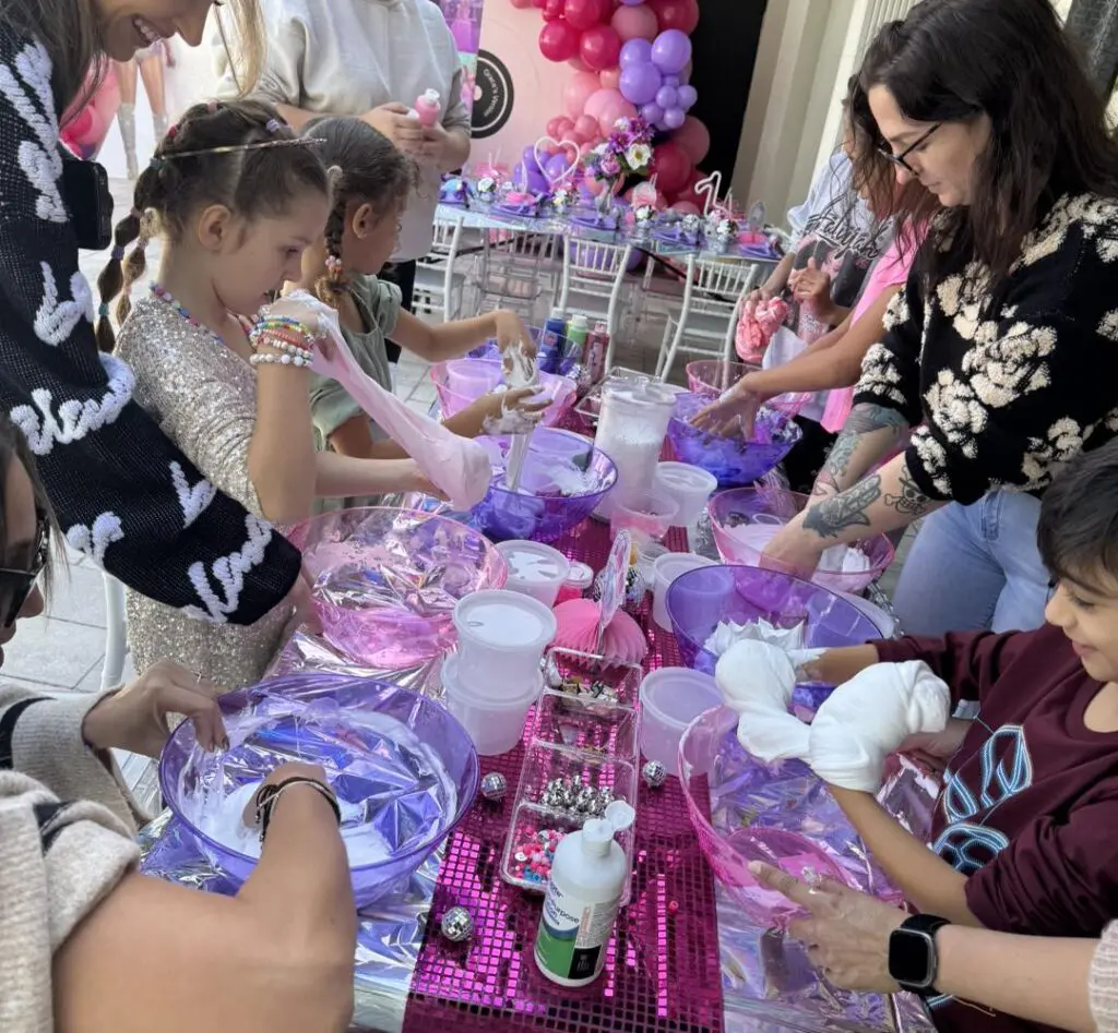 Children and adults immerse themselves in a fun-filled tie-dye activity at a colorful table brimming with vibrant dye bowls and artistic supplies. The scene is beautifully set against a lively backdrop featuring pink and purple decor, festive balloons, and the exciting atmosphere of renting a Movie Concession Stand. Perfect for capturing the joy of creative expression at any event, this engaging experience brings out smiles as participants transform plain white tees into unique works of art amidst an energetic celebration. Ideal for event planners seeking to add a splash of color and creativity to their gatherings!.