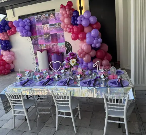 Experience the ultimate outdoor party setup in stunning cinematic style! A long table elegantly draped with a shimmering silver tablecloth serves as the centerpiece, complemented by rich purple napkins for a touch of sophistication. Lush floral centerpieces reminiscent of movie concession stand rentals add an artistic flair. The scene is brought to life by a vibrant balloon arch featuring lively shades of pink, purple, and red, creating the perfect backdrop for a memorable event under the sky. Perfect keywords: outdoor party setup, cinematic style, elegant table setting, floral centerpieces, vibrant balloon arch.