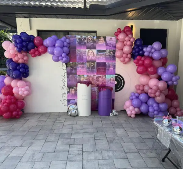 Colorful balloon decorations in vibrant shades of pink, purple, and red create a lively ambiance around an eye-catching backdrop featuring photos and a circular target design inspired by Movie Concession Stand Rental themes. The festive atmosphere is enhanced by two sleek cylindrical stands and a table brimming with delightful party favors, transforming the space into an inviting celebration hub. Perfect for event planning enthusiasts seeking creative balloon décor inspirations for memorable movie-themed parties!.