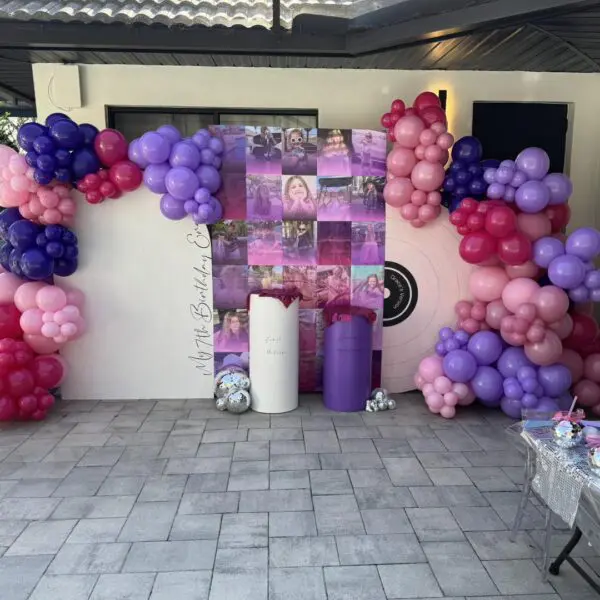 Colorful balloon decorations in vibrant shades of pink, purple, and red create a lively ambiance around an eye-catching backdrop featuring photos and a circular target design inspired by Movie Concession Stand Rental themes. The festive atmosphere is enhanced by two sleek cylindrical stands and a table brimming with delightful party favors, transforming the space into an inviting celebration hub. Perfect for event planning enthusiasts seeking creative balloon décor inspirations for memorable movie-themed parties!.