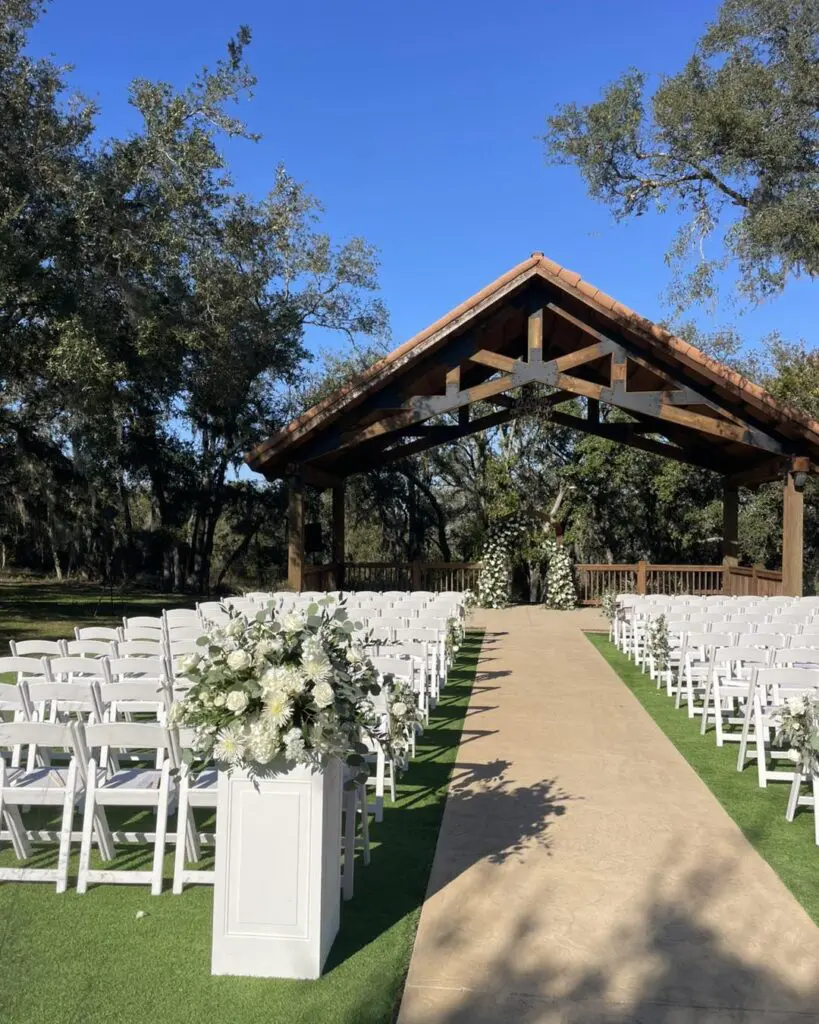 Elegant white chairs are meticulously arranged in two neat rows along a lush, green grassy aisle, guiding guests toward a beautifully crafted wooden pavilion. This picturesque setting features exquisite floral decorations that enhance its charm and sophistication. Nestled amidst a breathtaking backdrop of verdant trees, the scene exudes an inviting ambiance reminiscent of the enchanting allure of a vintage cinema's movie concession stand rental. Perfect for outdoor weddings or special events, this idyllic location marries nature's serenity with timeless elegance.