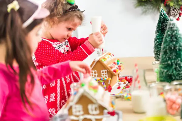 Two joyful children immerse themselves in the festive spirit as they craft gingerbread houses at a beautifully adorned holiday table. One child expertly handles a piping bag, adding intricate icing designs, while the other meticulously places colorful candy decorations. The scene captures warm holiday cheer and creativity, with gingerbread house decorating ideas and family bonding at its heart.