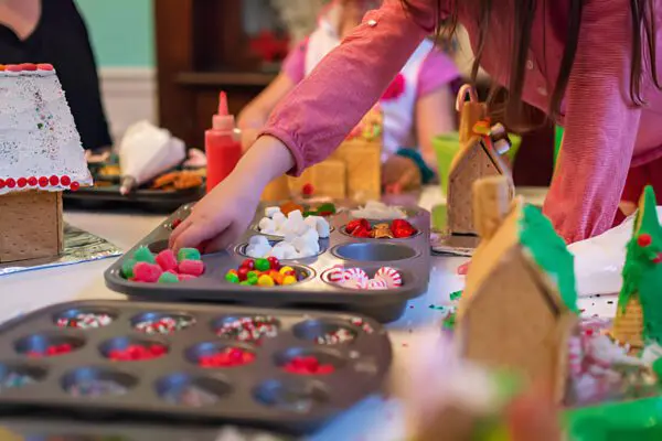 Children joyfully decorating festive gingerbread houses with an array of colorful candies and icing. A hand eagerly reaches for vibrant sweets nestled in a muffin tin, adding to the creative culinary masterpiece. Perfect activity for holiday fun and family bonding, ideal for Christmas crafts with kids.