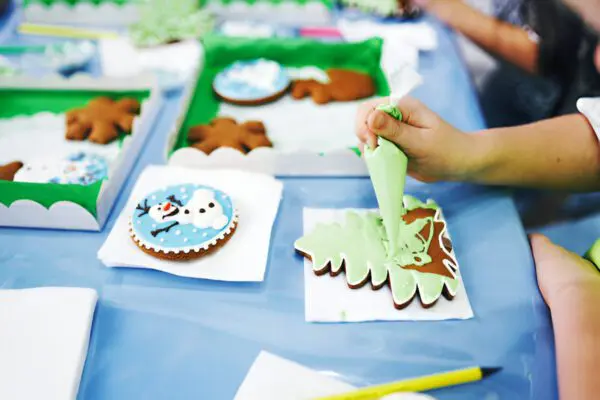 Festive cookie decorating session at a table filled with colorful icing tubes and holiday-shaped cookies. A participant skillfully applies vibrant green icing to a Christmas tree-shaped cookie, surrounded by an array of sweet treats ready to be adorned. Perfect for DIY holiday baking and creative dessert decoration ideas.