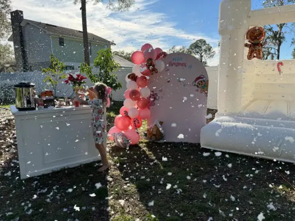 In a magical outdoor winter wonderland, a joyful child gleefully plays amidst vibrant pink and red balloons. The scene is beautifully set with a grand white bouncy castle that stands as a centerpiece, adorned with twinkling decorations resembling glistening snowflakes, courtesy of the Snow Machine Rental. A festive sign adds to the ambiance with its cheerful greeting: "Merry Christmas." Perfect for holiday celebrations, this enchanting setting captures the spirit of Christmas joy and fun.