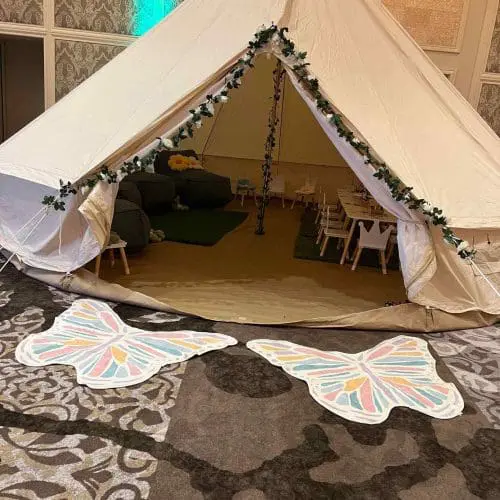 A large indoor tent decorated with leafy garlands at the entrance, flanked by two butterfly-shaped mats on a patterned carpet, perfect for various party themes.