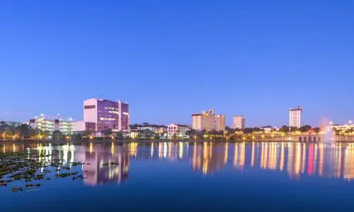 Lakeland party rentals. City skyline at twilight with modern buildings reflected in a calm lake, under a clear blue sky in the target locations.