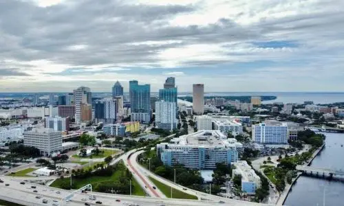 Areas We Serve: Aerial view of a city skyline with tall buildings near a body of water, cloudy skies above, and roads weaving through the landscape.