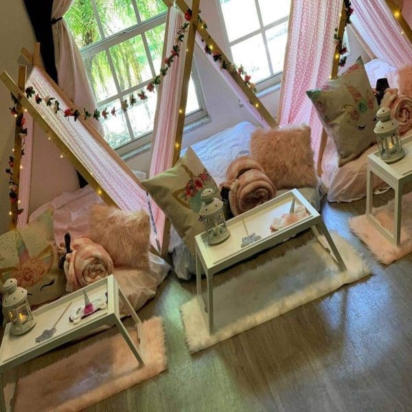 Three indoor teepee tents with pink decor, two pillows each, surrounding white tables. Blankets and lanterns are placed on the tables, perfect for a cozy slumber party. The window in the background shows greenery outside. Ideal for a charming party rental setup.