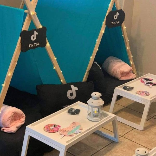 Two indoor teepees with TikTok logos, blue covers, and fairy lights are positioned on the floor with white trays in front, each holding eye masks and small decorative items. Pink blankets and pillows inside set the scene for a cozy slumber party or a fun-filled slime party.
