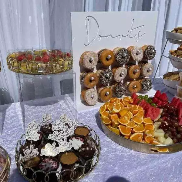 A dessert table adorned with a donut wall, cupcakes, assorted fruits, and various desserts displayed in gold-rimmed trays is perfect for any event. Complement this setup with our XL Outdoor Games Addon to create an unforgettable experience. Our party service ensures everything looks impeccable, making it an ideal choice for kids' party rentals or any celebration requiring top-notch party rental options.