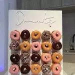 A display stand holding four rows of assorted donuts, including chocolate, pink frosted, plain glazed, and sprinkles, arranged neatly in a 5x4 grid pattern. Perfect for your next slumber party or event from our XL Outdoor Games Addon rental service. The word "Donuts" is written at the top.