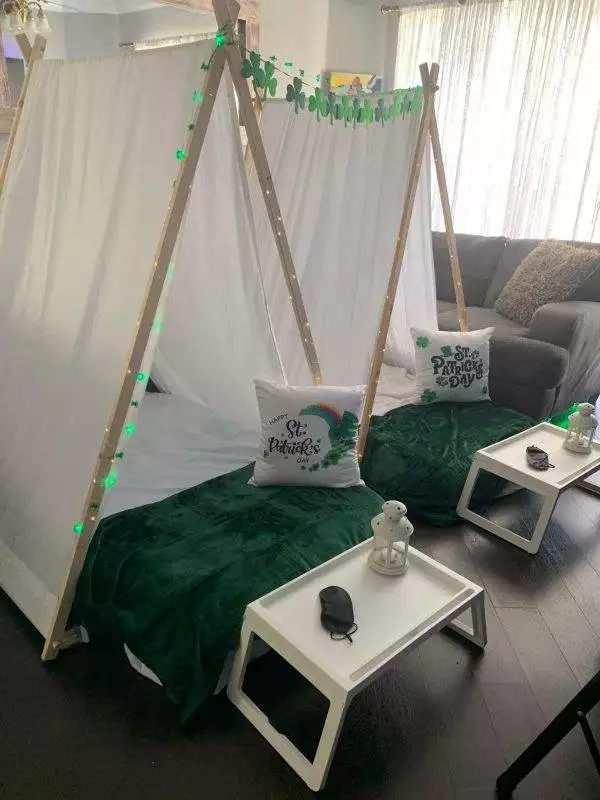 Two makeshift tents, perfect for a kids' party rental, are decorated for St. Patrick's Day with white sheets and green blankets. Each teepee features themed pillows, string lights, and a small tray table with a lantern and face masks.