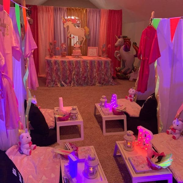 A room decorated for a unicorn-themed slumber party with pink and purple drapes, unicorn balloons, plush unicorn toys, colorful banners, small tables with lights, and seating cushions on the floor. Perfect for a kids party rental!
