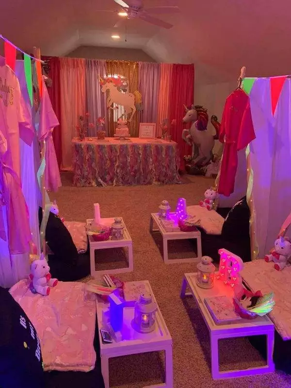 A room decorated for a unicorn-themed slumber party with pink and purple drapes, unicorn balloons, plush unicorn toys, colorful banners, small tables with lights, and seating cushions on the floor. Perfect for a kids party rental!