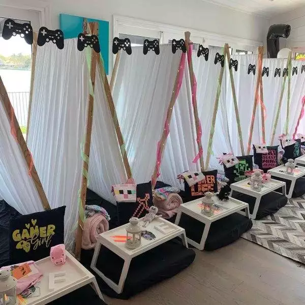 Indoor tent setup with gaming-themed decorations, including black and pink pillows, gamer-themed pillowcases, and controller cutouts above each tent. Perfect for a kids party rental, small trays with snacks and drinks are in front to keep everyone fueled for fun.