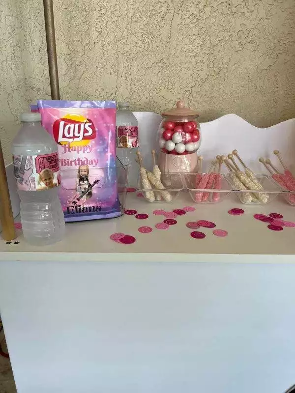 A decorated table for a kids party rental features a birthday Lay's chips bag, water bottles, gumballs in a jar, and candy sticks. A festive touch is added to the table's surface with pink and white confetti scattered around. The setup is completed with a Large Greenery Photo Backdrop featuring a vibrant neon sign.