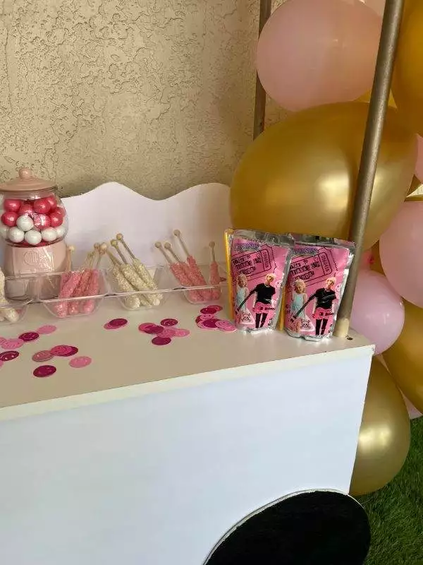 A Large Greenery Photo Backdrop with Neon Sign, adorned with cake pops, gum balls, two packs of pink-themed juice, and pink and gold balloons on the side, is perfect for a kids party rental. Pink and black round confetti is scattered around for added fun.