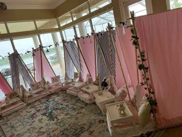 A room setup with pink and striped fabric teepees, cushions, and low tables, designed for a slumber party event, overlooks a scenic lake view through tall windows.