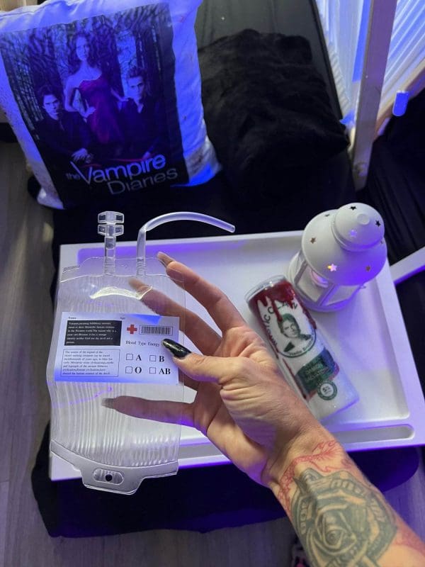 A hand holds a blood bag drink container with a blood type checklist. In the background, there's "The Vampire Diaries" pillow, a white lantern, and a novelty bag with a "Forest Queen" design, all part of an enchanting teepee party rental setup perfect for your next themed event.