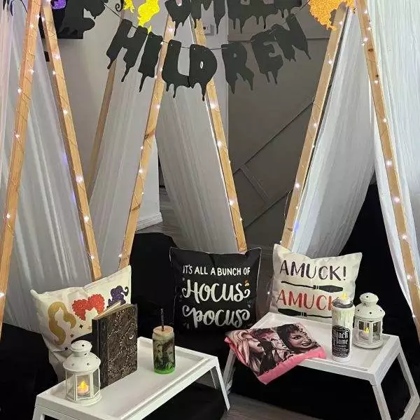 Two wooden tents are decorated with Halloween-themed items and strung lights, perfect for a kids party rental. Each tent has side tables with lanterns and candles, with pillows featuring spooky designs and phrases like "It's all a bunch of Hocus Pocus.