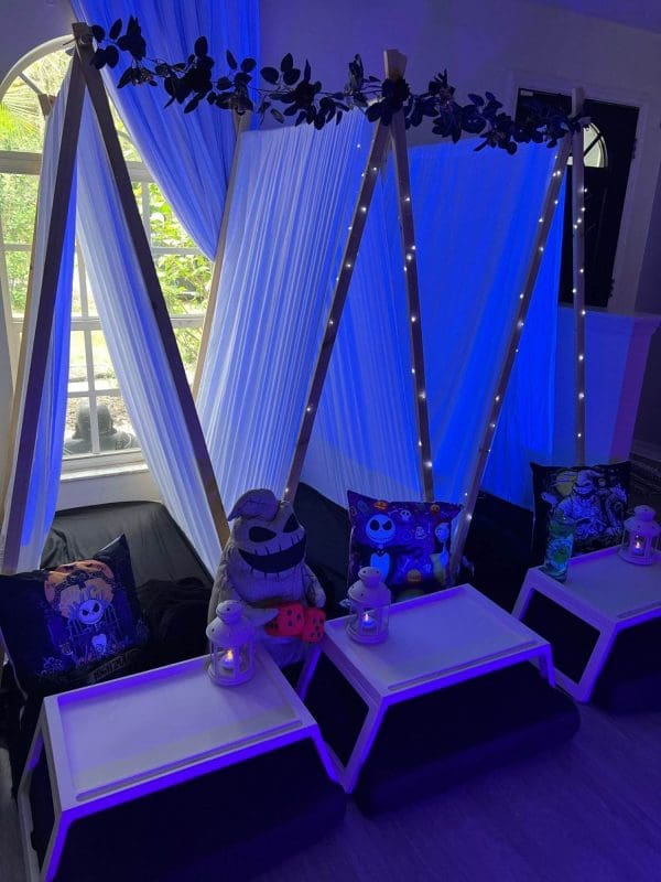 A cozy indoor setup with teepee tents, blue lighting, white curtains, and Halloween-themed pillows and decorations creates the perfect ambiance. Small tables with lanterns are positioned in front of each tent, ideal for a slime party or any fun-filled party rental occasion.