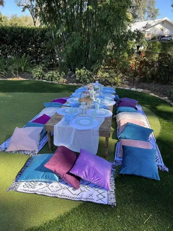 An outdoor picnic setup with a long wooden table covered in white cloth, set for a meal, surrounded by colorful cushions on a neatly mowed lawn—perfect for a kids party rental.